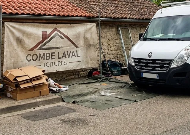 Bâche Combe Laval Toitures tendue devant un mur en pierre à Saint-Jean-en-Royans, cartons et bâche de sol rangés, fourgon garé le long du chantier