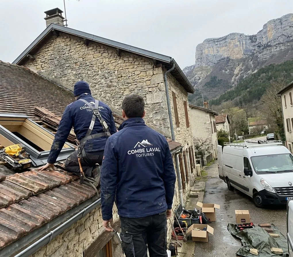 Deux couvreurs Combe Laval Toitures, dont un encordé, posent une fenêtre de toit sur une toiture en tuiles à Saint-Jean-en-Royans, falaises du Vercors au fond